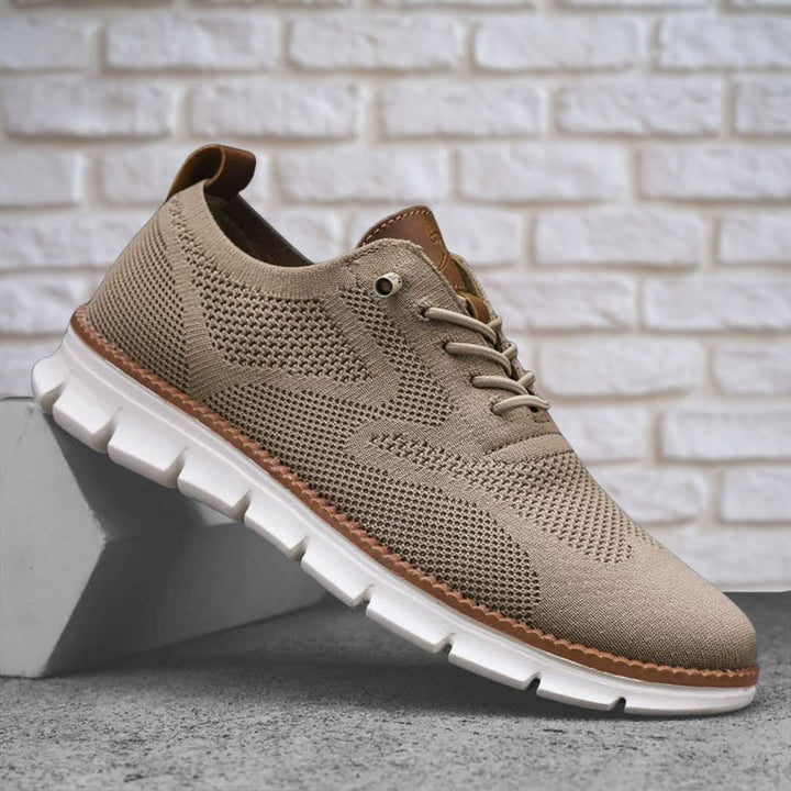 Taupe knit trainer with a white sole resting on a block against a white brick wall