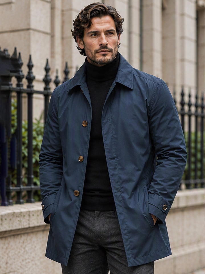 Men's trench coat in navy, a single breasted mid length mac coat worn over a black roll neck on a city street